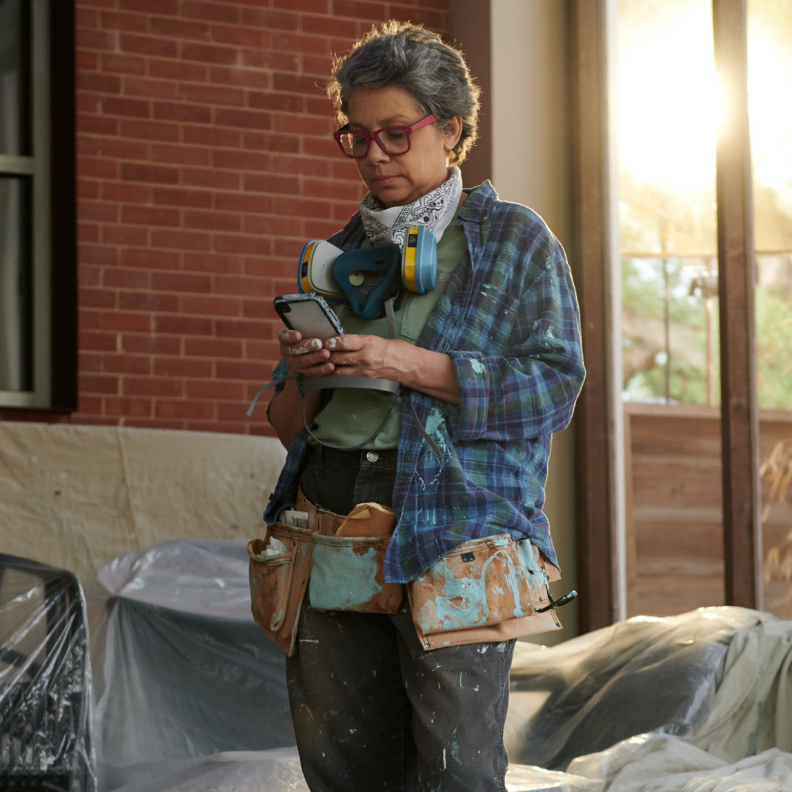 Painter looking at her phone. Wears red glasses, flannel shirt and tool belt, with respirator and bandana around neck. 