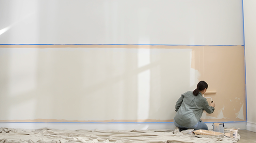 Woman kneeling in front of large taped-off section of wall, using large roller to apply a neutral beige.