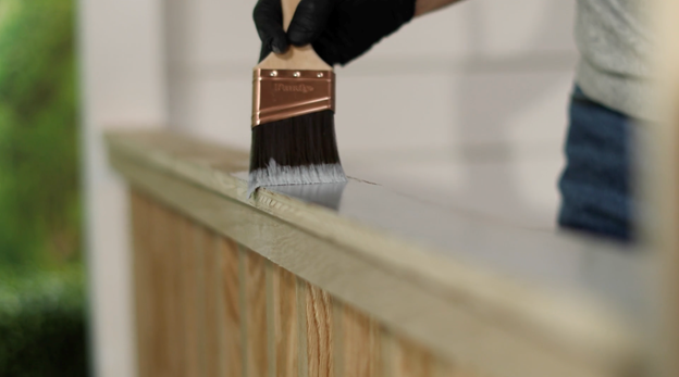 Gloved hand with brush applying stain to wood railing.