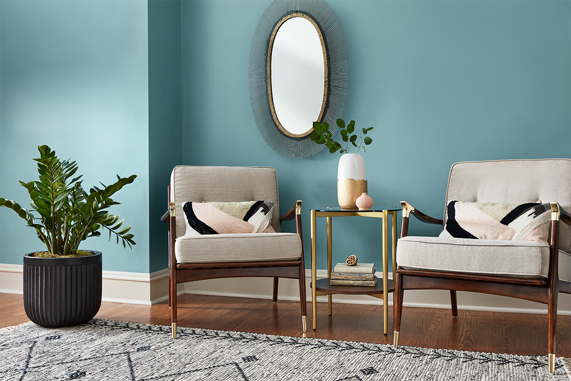 Matching wood-and-brass armchairs with brass-and-glass table set against cool blue wall with oval mirror.