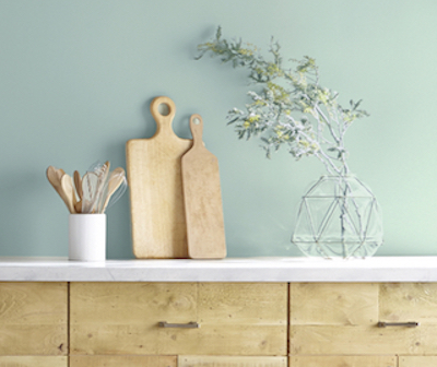 Light, sage-green kitchen walls, wood cabinets with white countertop, cutting boards, vase and spoons.