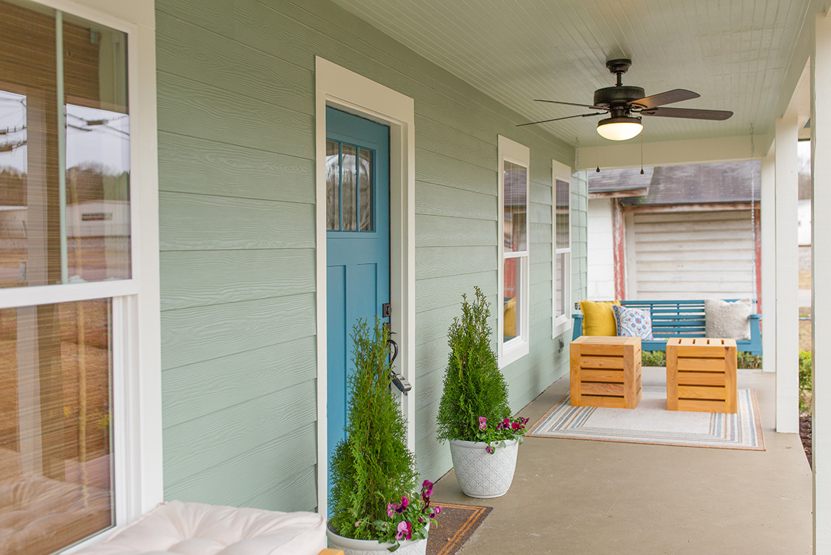 View along porch of light-green house, blue door with potted evergreens, two square tables and blue porch swing.