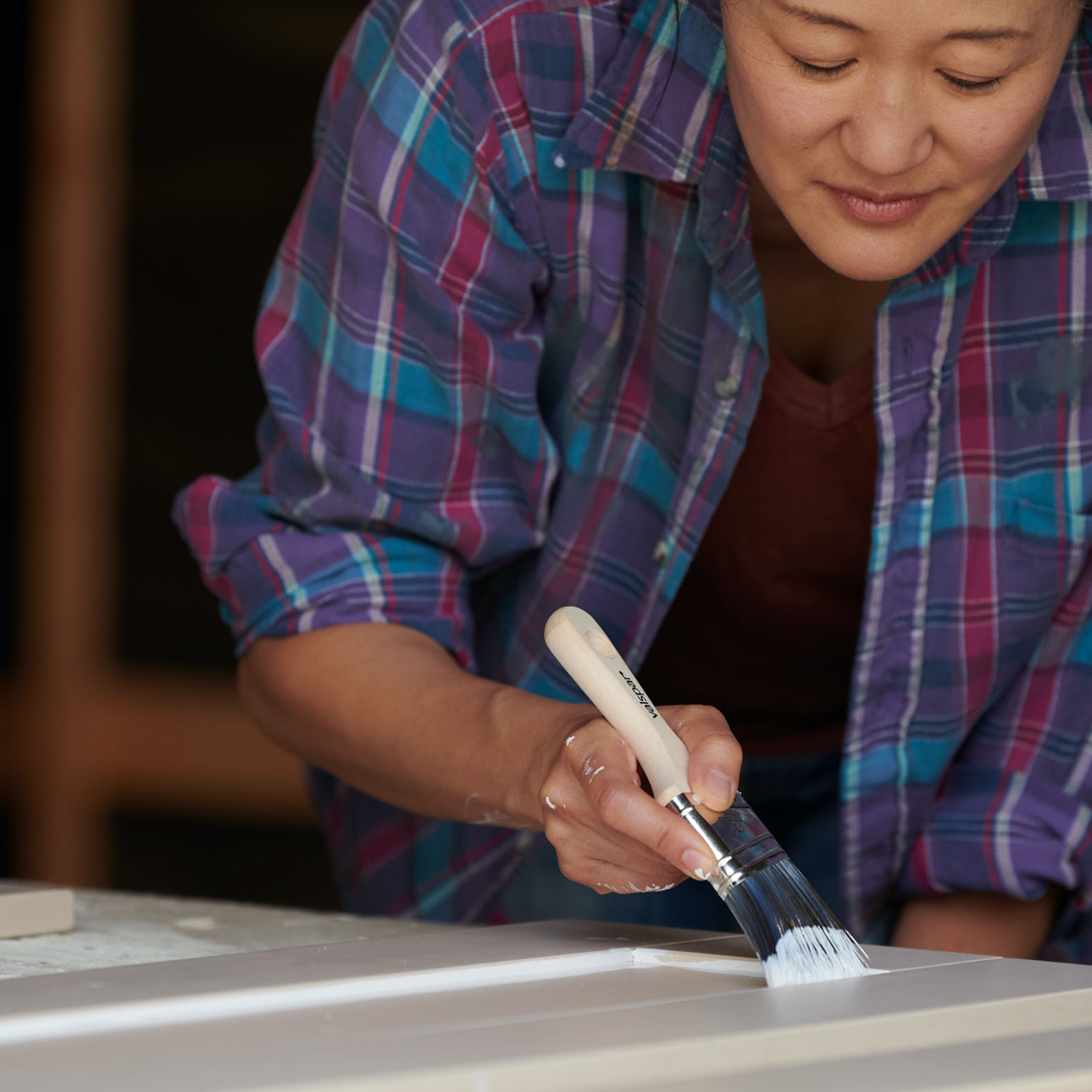 Woman with brush painting cabinet doors which are laying on a table.