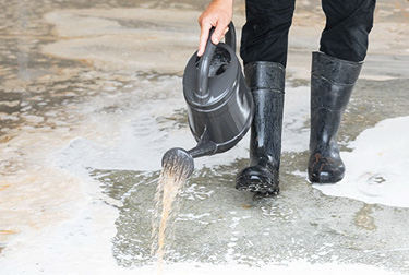 Person in rubber boots with watering can clearing liquid off concrete by pouring water from the can. 