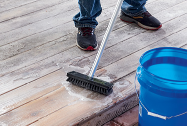 Person in jeans and sneakers using push brush to distribute liquid across a wood surface.