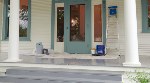 The front porch of a home where painting is in process. A drop cloth, ladder and other painting tools are on the porch.