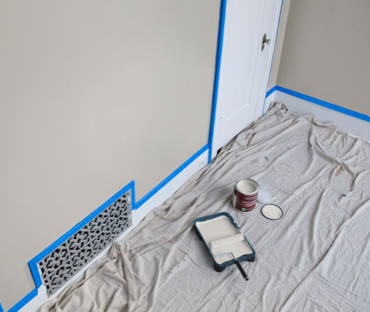 Drop cloth next to masked off wall. Off-white paint and roller in tray. Open can of paint with lid off. 