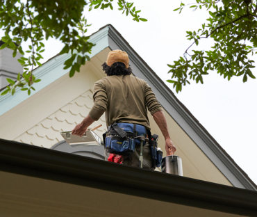 Painter holding paint can, tray and brush looks up to peak of roof with creamy neutral scallop siding and gray trim.