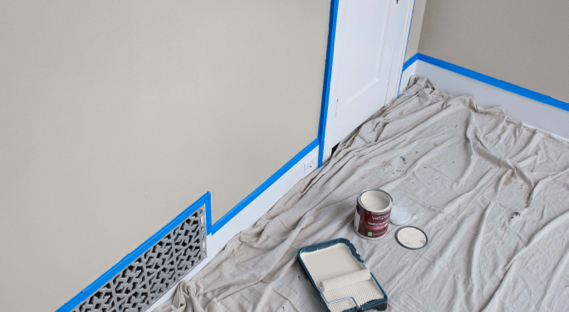 Drop cloth next to masked-off wall. Off-white paint and roller in tray. Open can of paint with lid off. 