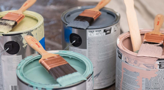 Four open paint cans with brushes and stirrers laying across the tops: soft yellow, dark gray, blue-green and pink.