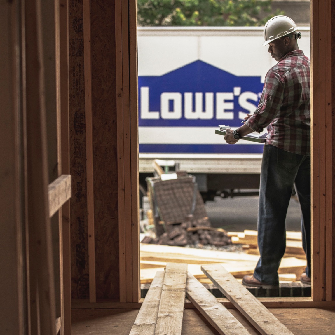 Imagen de un trabajador de la construcción mirando un portapapeles en medio del suelo y un camión de Lowe's en el fondo