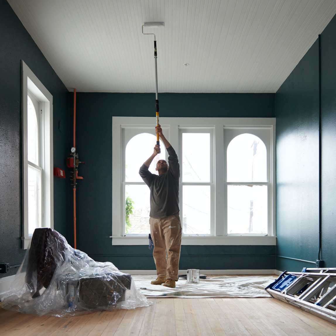 Person in a large room with windows using a roller on an extension pole to paint the ceiling.
