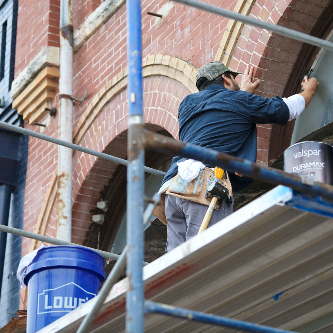 Man on a scaffold in front of a brick building painting window frame with a blue bucket and can of paint in the foreground.