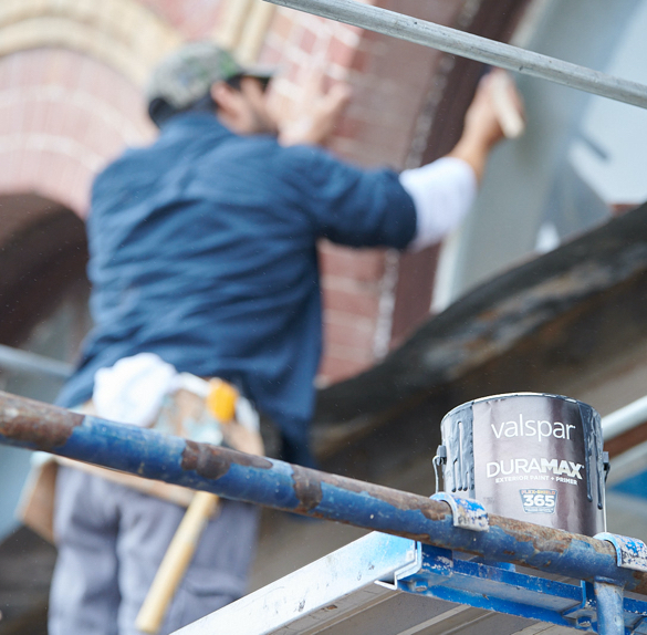 Person on a scaffold in front of a brick building painting window frame with a can of paint in the foreground.