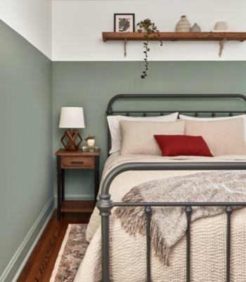 Bedroom with lower half of wall in Secret Moss and upper half in a neutral white. Iron-post bed with bright red pillow.