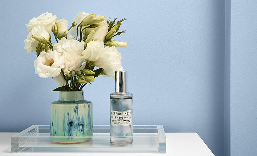 Close-up of flowers in a vase and a perfume spray in a clear tray on a bathroom counter. Walls are light blue. 