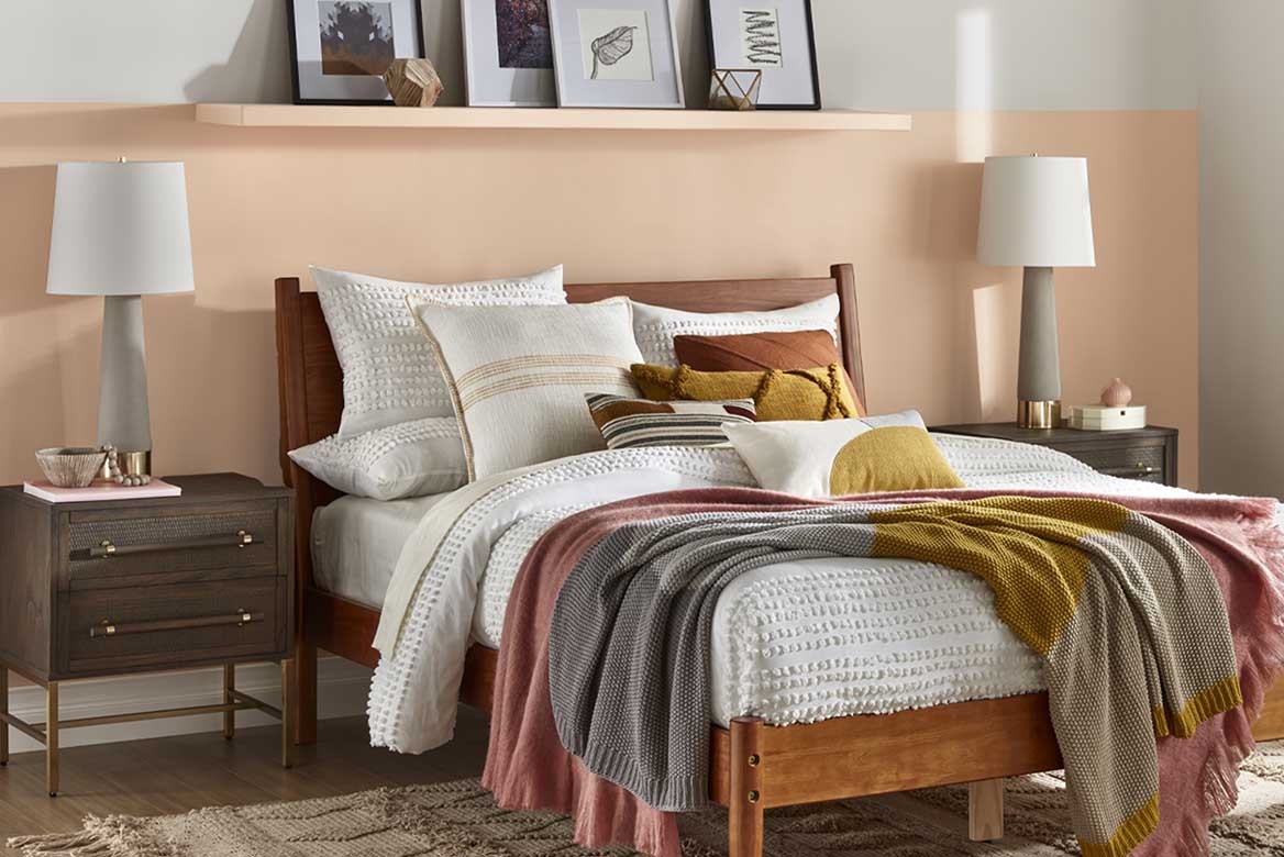 Pillow-covered bed against sandy neutral half wall accent color with matching shelf and modern side-table lamps.