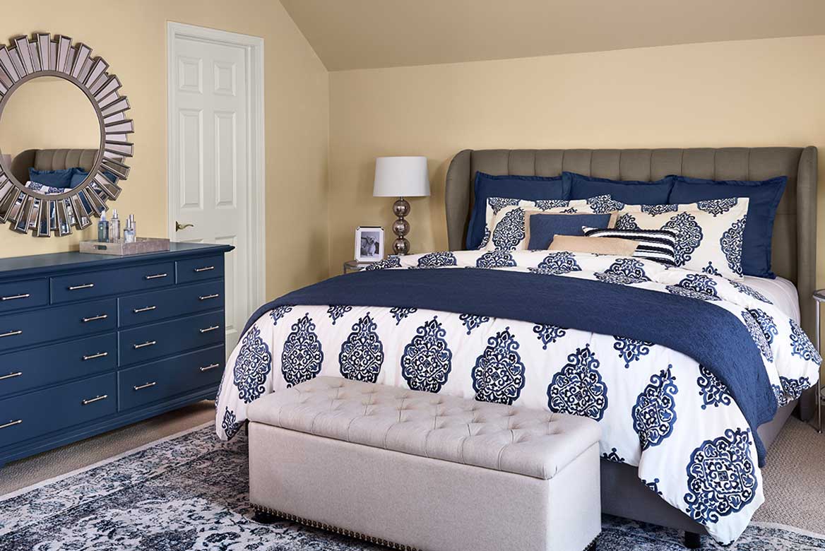 Warm neutral bedroom walls with blue dresser and upholstered bed with coverings in shades of patterned blue and white.
