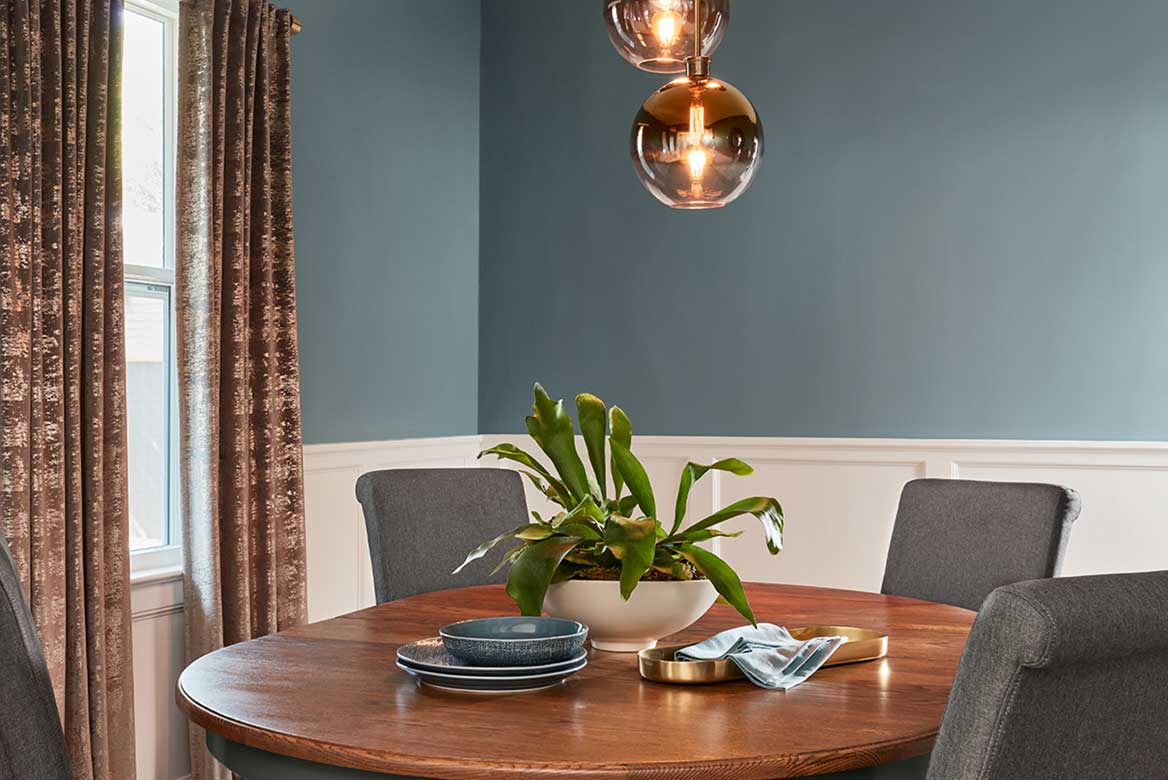 Small dining room with bluish gray walls and half wall panels, oval wood table and hanging globe lights.