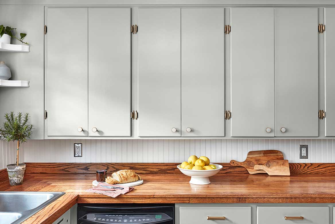 Mid-tone gray upper kitchen cabinets above wood countertop upon which sit fresh bread and raised bowl of lemons.