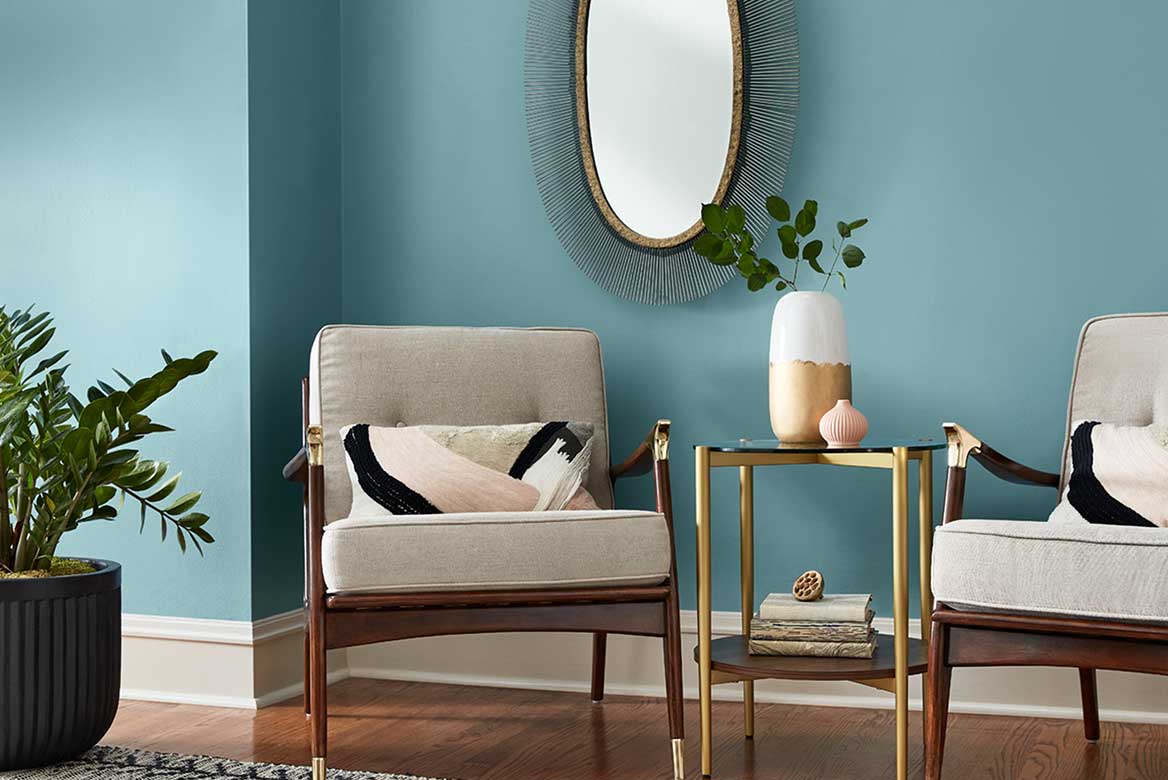 Matching wood-and-brass armchairs with brass-and-glass table set against cool blue wall with oval mirror.