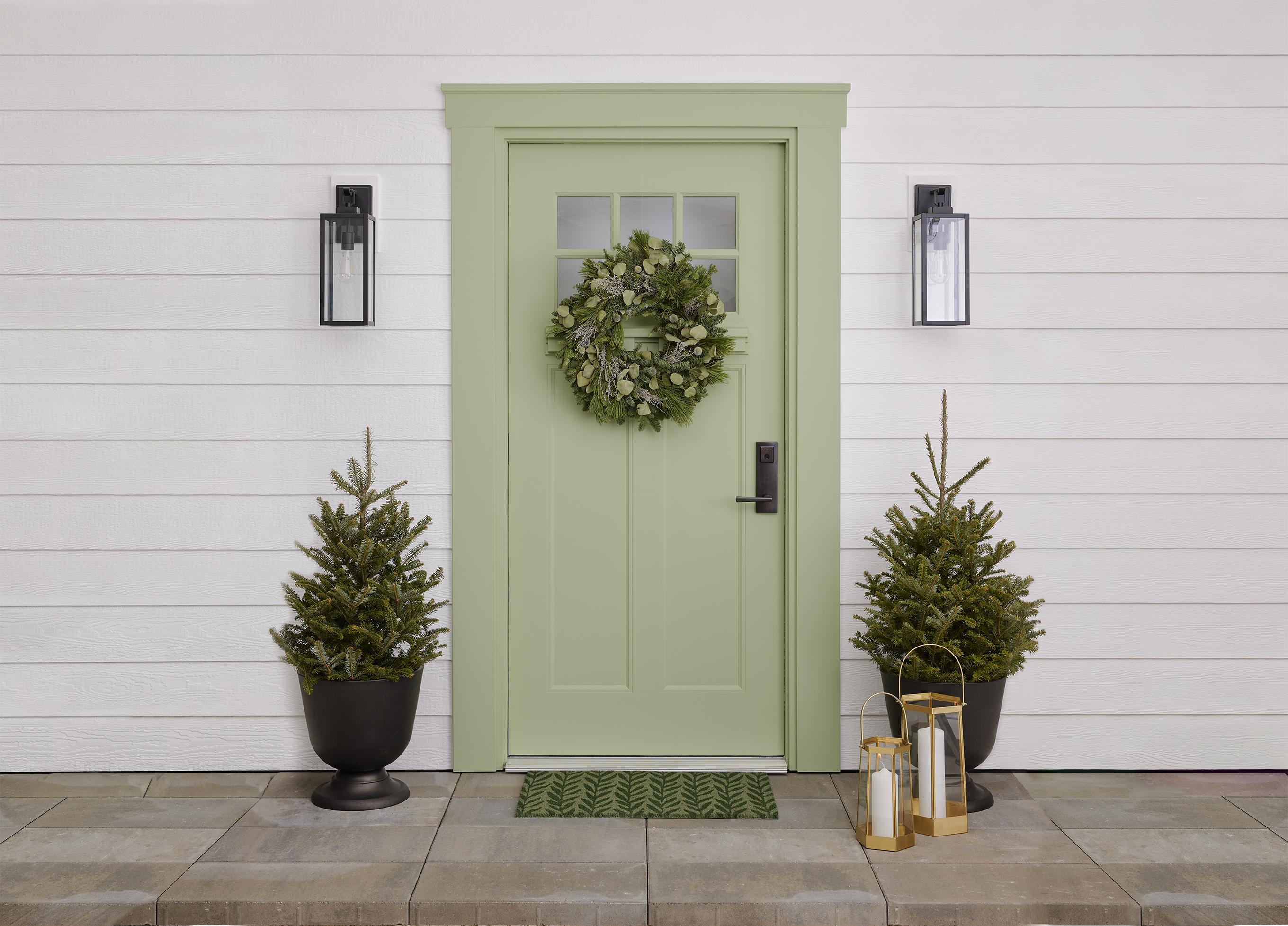 Pale-green door with wreath and doormat. Two potted evergreens and gold lanterns frame the door.