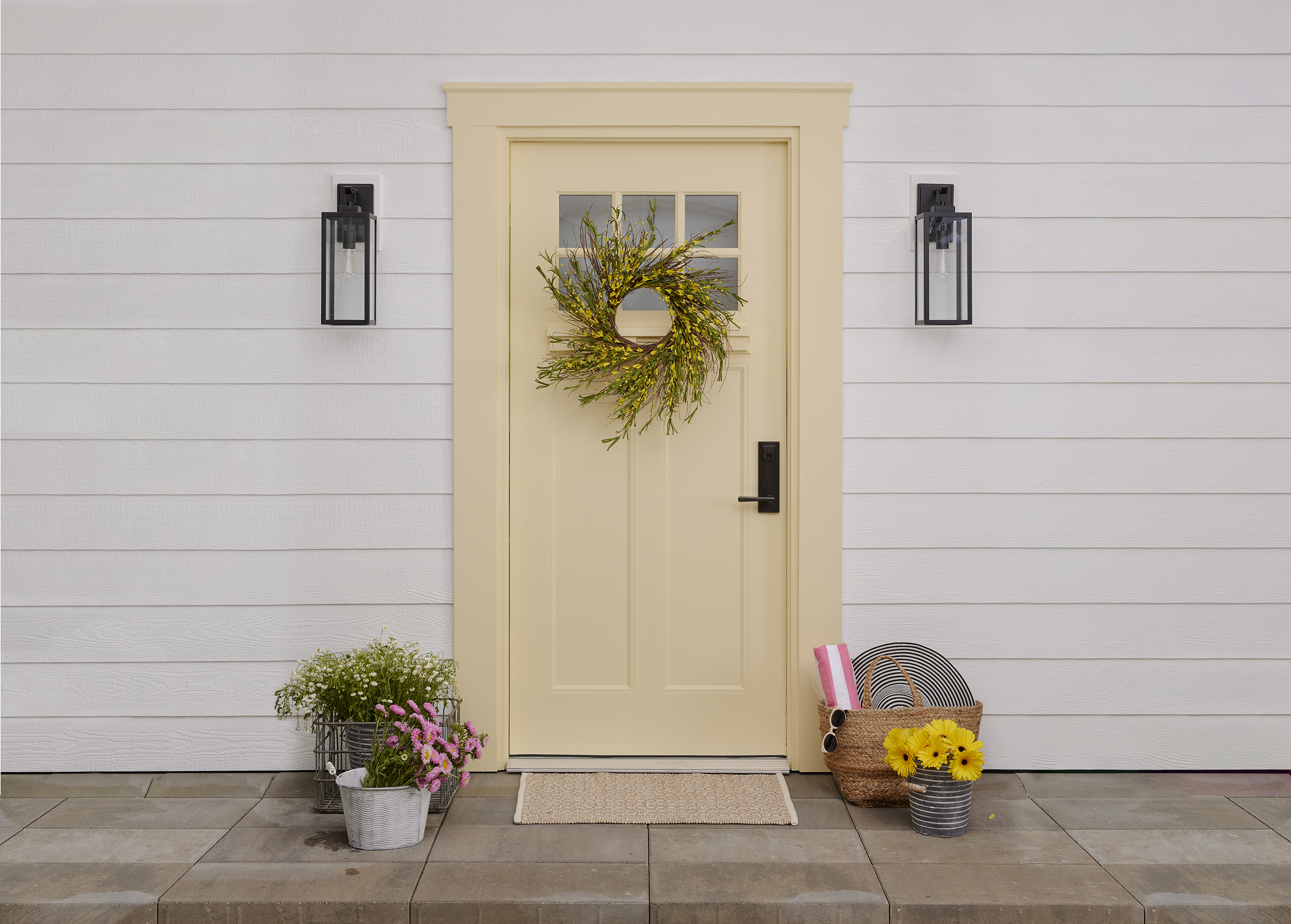 A buttery yellow door with a wreath and doormat. A beach basket and containers with fresh-cut flowers rest near the door.