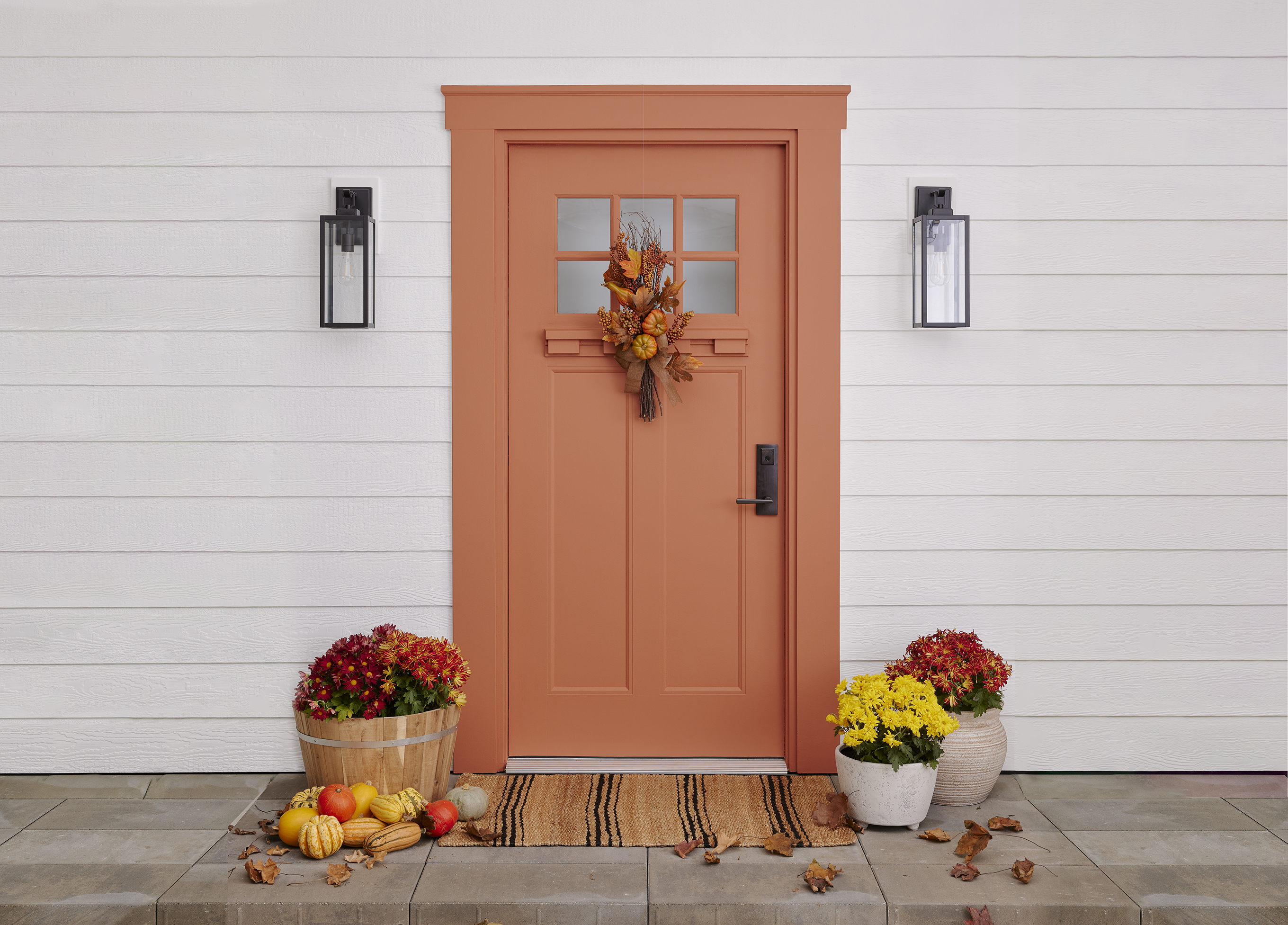 A clay-red door with autumn wreath and doormat with potted plants in fall shades, fall leaves and colorful gourds.