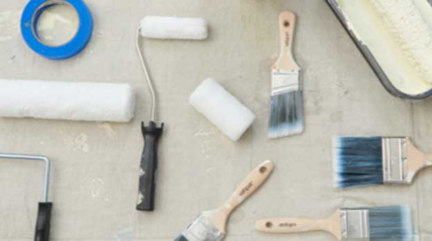 Used brushes and rollers laying in different directions on a paint-stained drop cloth.