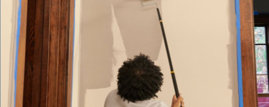 Back of person using extension pole to apply a brownish-tan color to a wall with taped off trim.