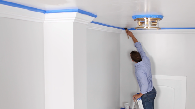 Person on ladder using a brush to apply white color to ceiling with blue tape visible around the trim.