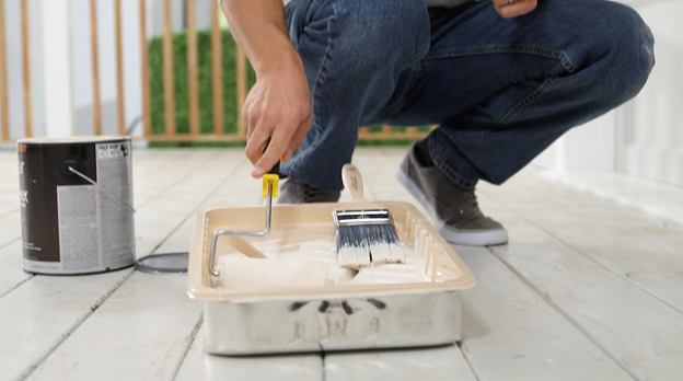Bottom half of person on unfinished patio using a roller tray containing a brush and an open can in the background.