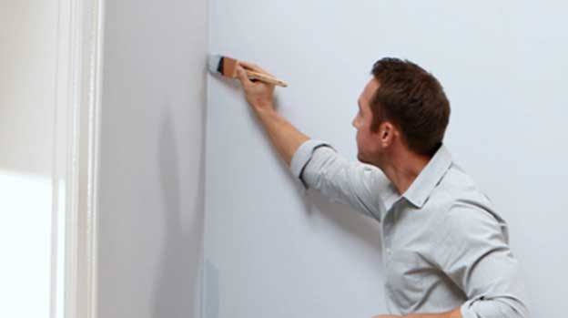 Person leaning forward with brush in hand applying paint to the corner of a wall.