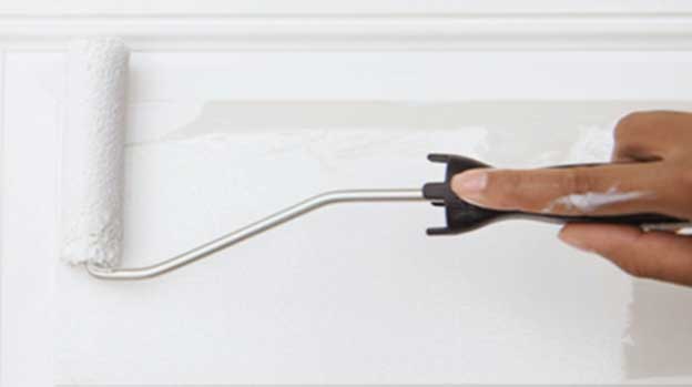 Roller applying white paint to wall with person's index finger on handle.