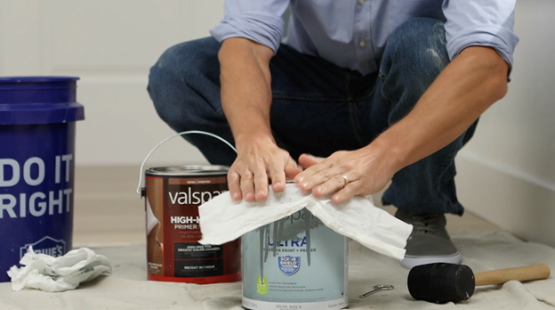 Man squats on drop cloth behind Valspar Ultra paint can, using rag to wipe rim. Rubber hammer and paint cans in background.