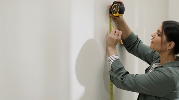 Woman holds measuring tape and uses a pencil to mark where a shelf should be placed on a wall.
