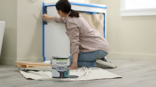 Out-of-focus woman painting a prepped and taped vanity. Can of Valspar Cabinet & Furniture paint in focus in foreground.