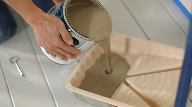 Hands pouring brownish-tan color into a tray with paint can opener on floor.
