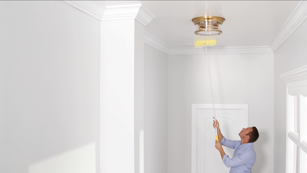 Person in front of a white doorway. using sponge with extendable handle to clean ceiling around a gold light fixture.