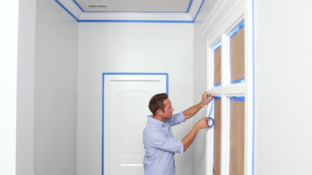Person applying blue painter's tape to windows, doorway and ceiling.