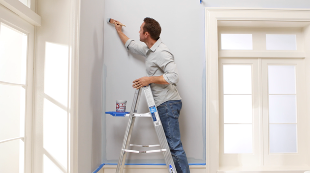 Person on ladder using a brush to apply white color to ceiling with blue tape visible around the edges.