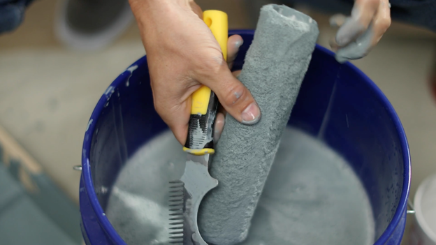 Close up of bucket filled with gray color with hand holding a tool and used roller cover.