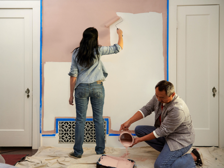 Person using roller to apply color to wall between two doors with another person pouring a can into tray.