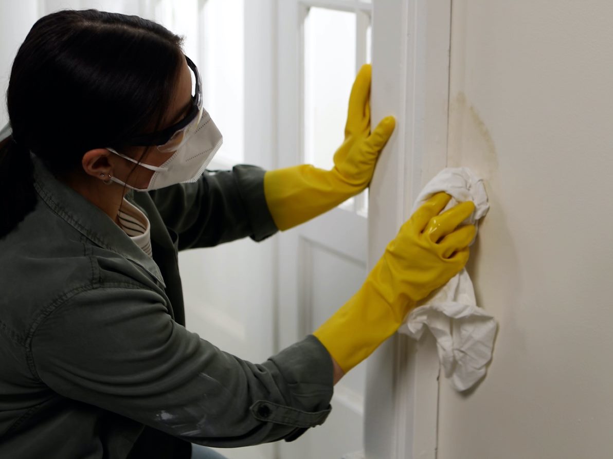 Person with safety glasses, mask and yellow rubber gloves scrubbing stain off wall.